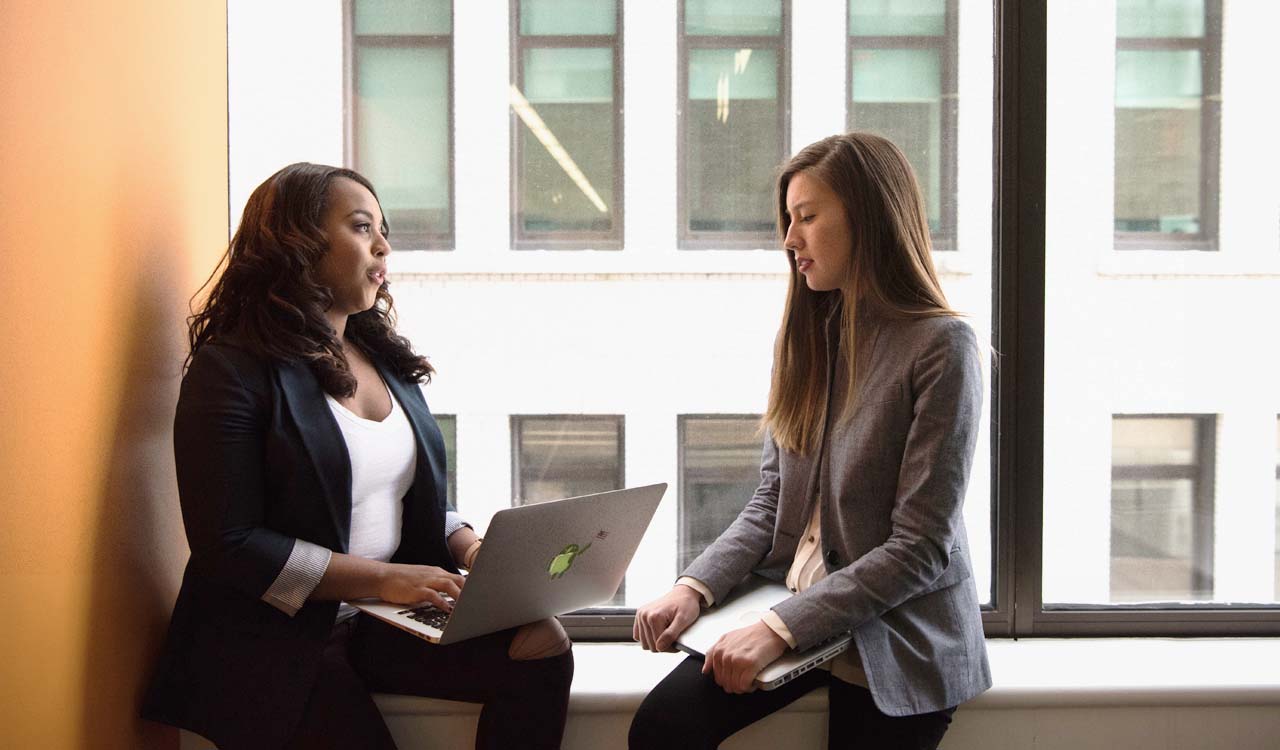 Two IT professionals sitting on a windowsill holding laptops and talking.