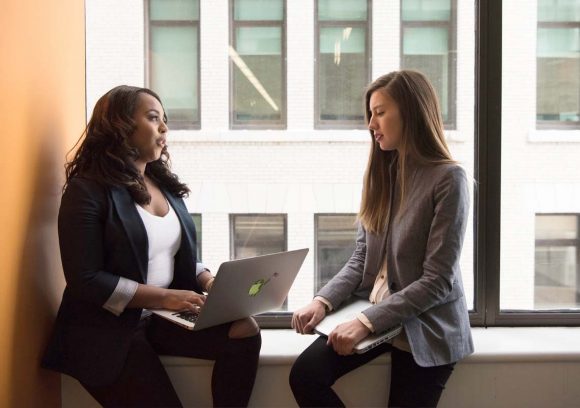 Two IT professionals sitting on a windowsill holding laptops and talking.