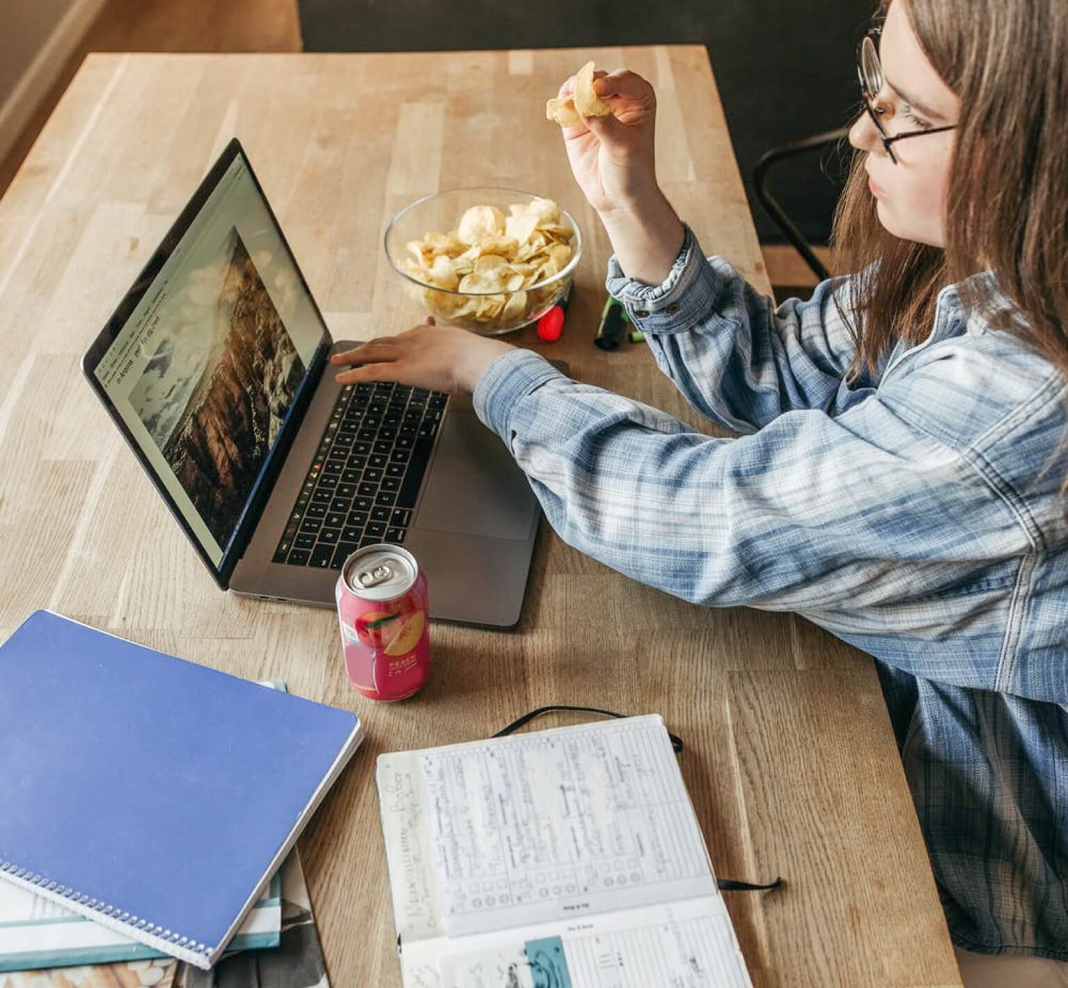 A person studying on a table with books and a laptop while eating a snack.