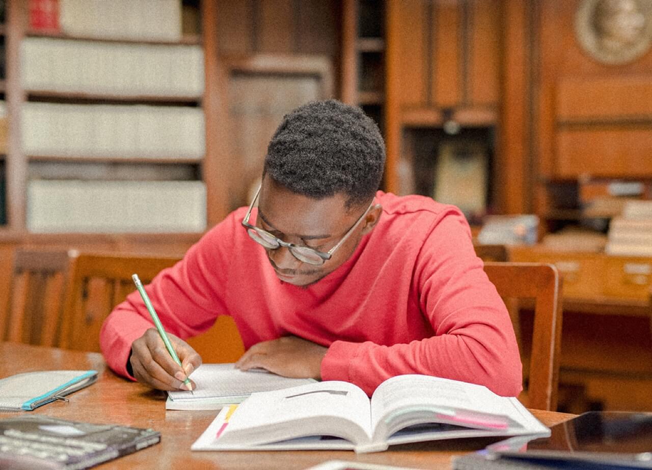 A person studies at a desk taking notes while bent over books.