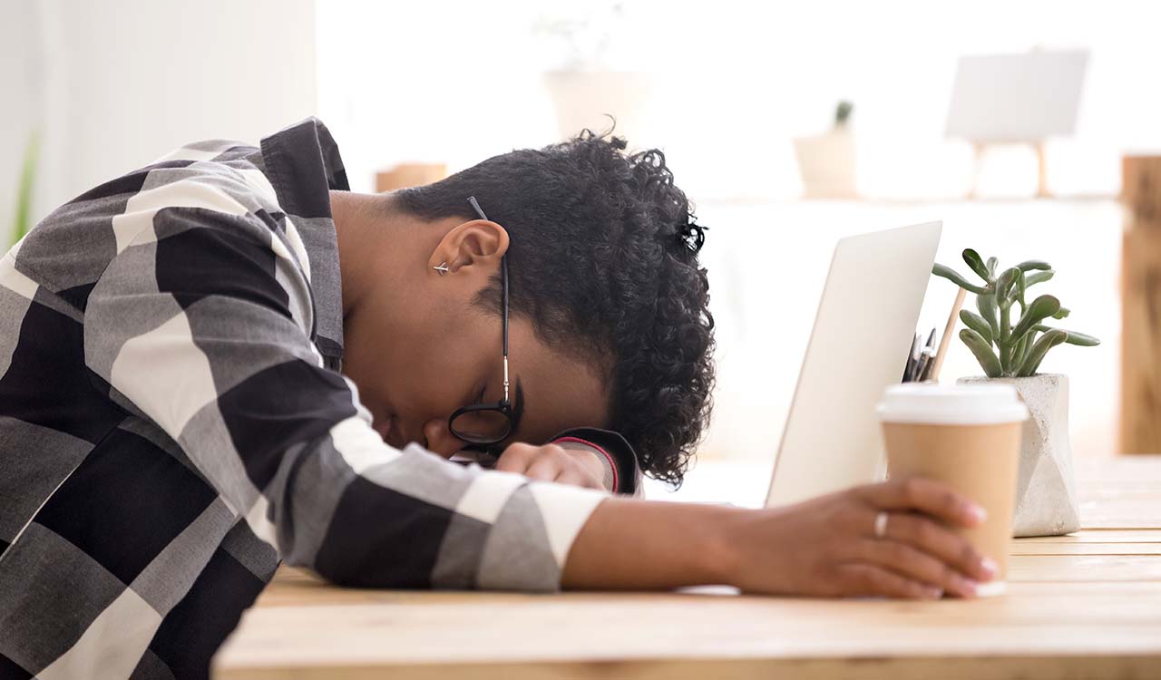 A frustrated person with their head down on top of a computer while holding coffee.