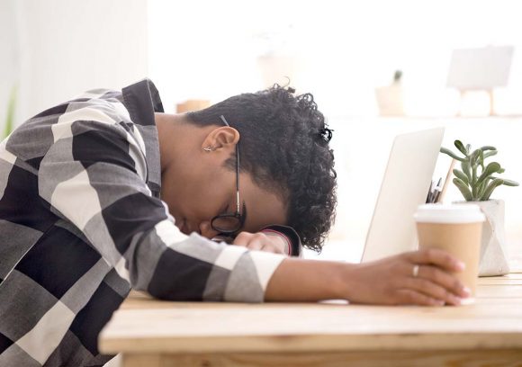 A frustrated person with their head down on top of a computer while holding coffee.