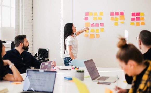 A project manager stands at a white board and organizes post it notes for a team.