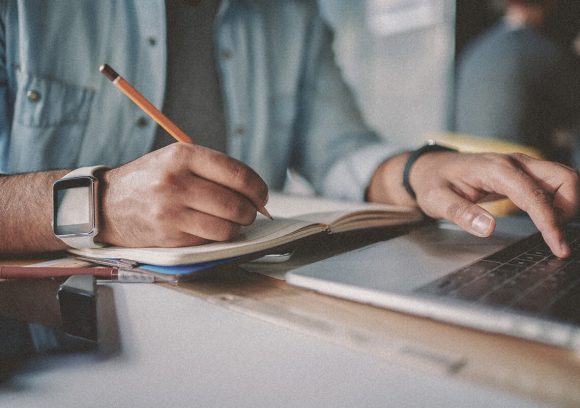 A person studying on a computer and taking notes in a note book. Closeup of hands.