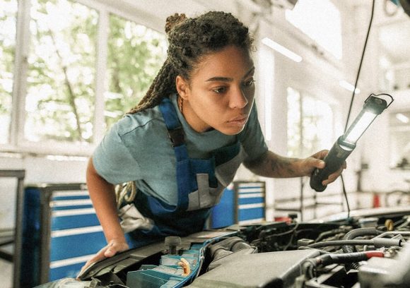 An automotive technician inspects a car engine while holding a light.