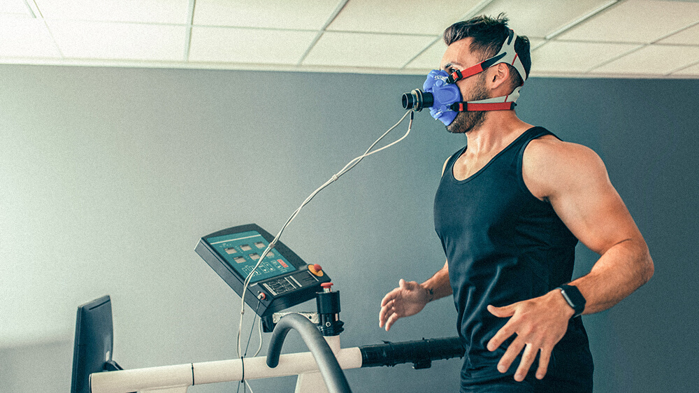 Man running on treadmill wearing oxygen mask attached to a monitor.