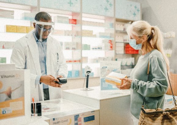 A pharmacist interacts with a patient behind a plexiglass barrier.