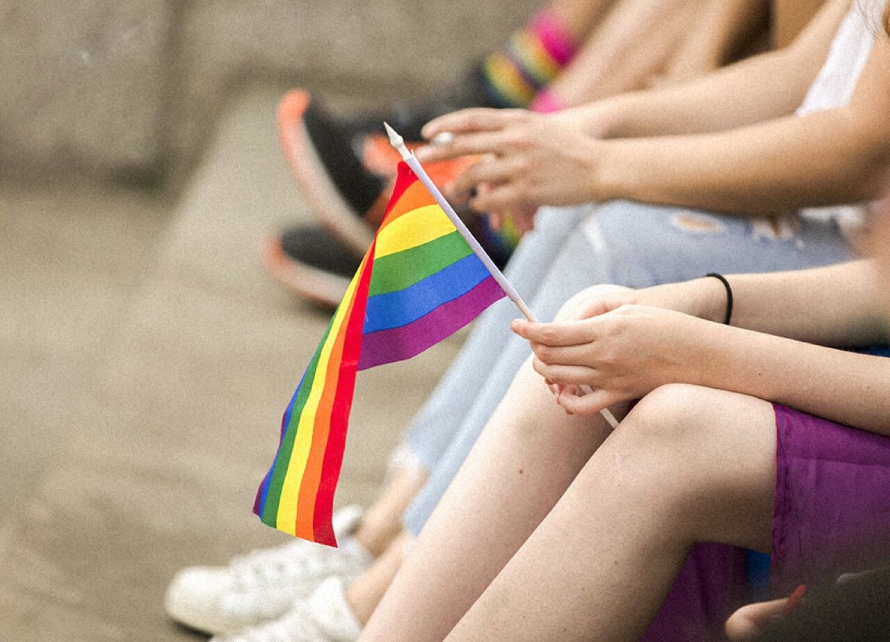 A group of young people sitting on steps while one holds a rainbow flag. Close-up of legs and hands.