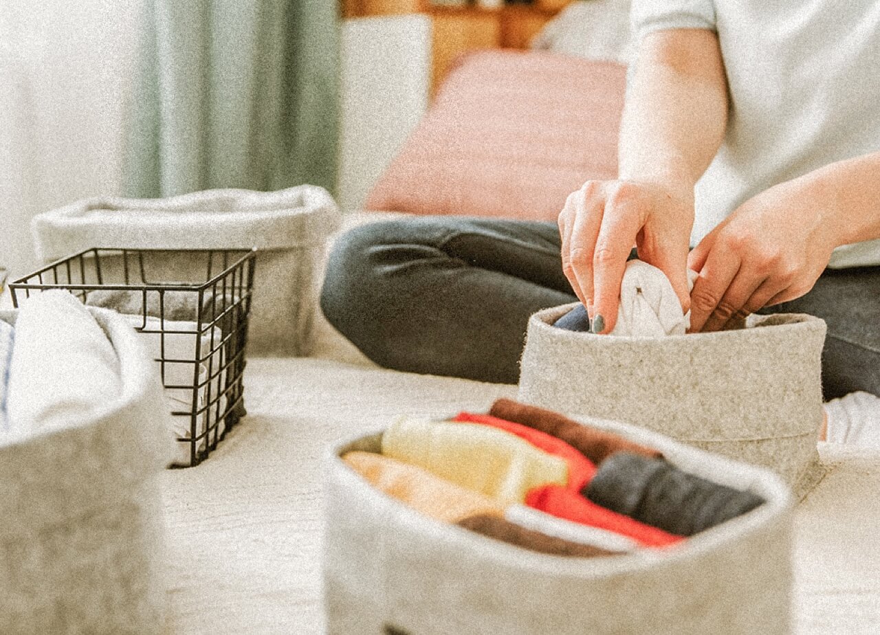 A person sits on the floor folding clothes and neatly placing them into fabric storage bins.