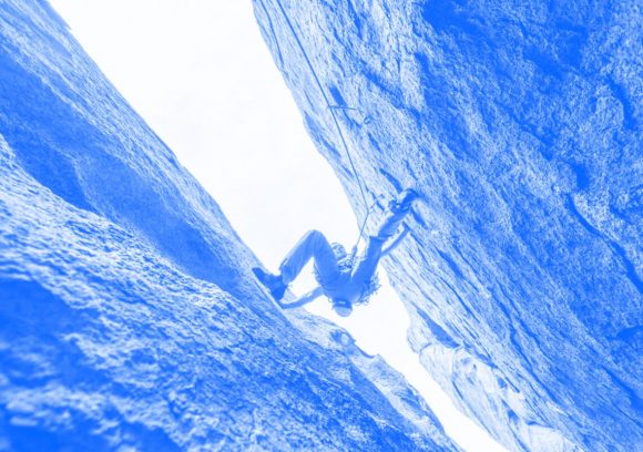 Underneath view of a rock climber scaling between two rock crevices. Blue-toned.