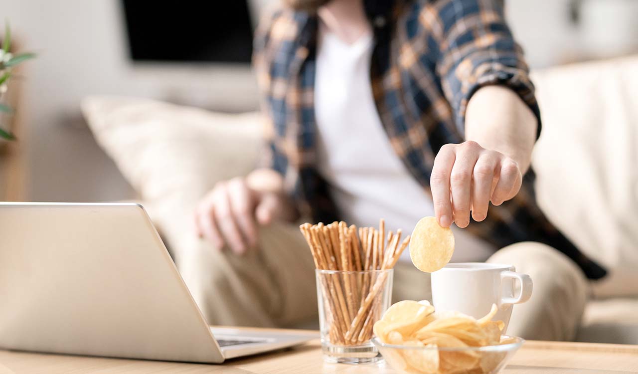 A person sitting on a couch reaches to eat some chips from a pile of snacks on a coffee table.