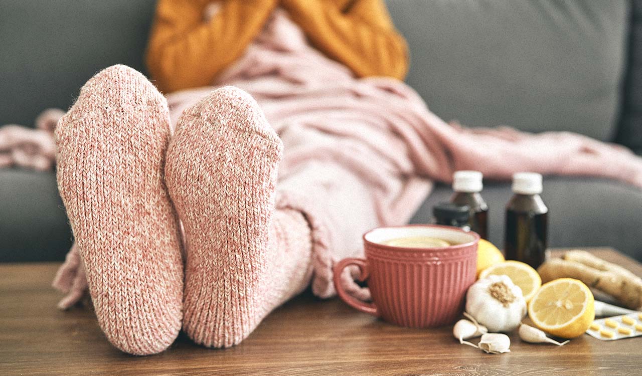 A coffee table with home cold remedies like tea, garlic and ginger with a person's socked feet resting nearby.