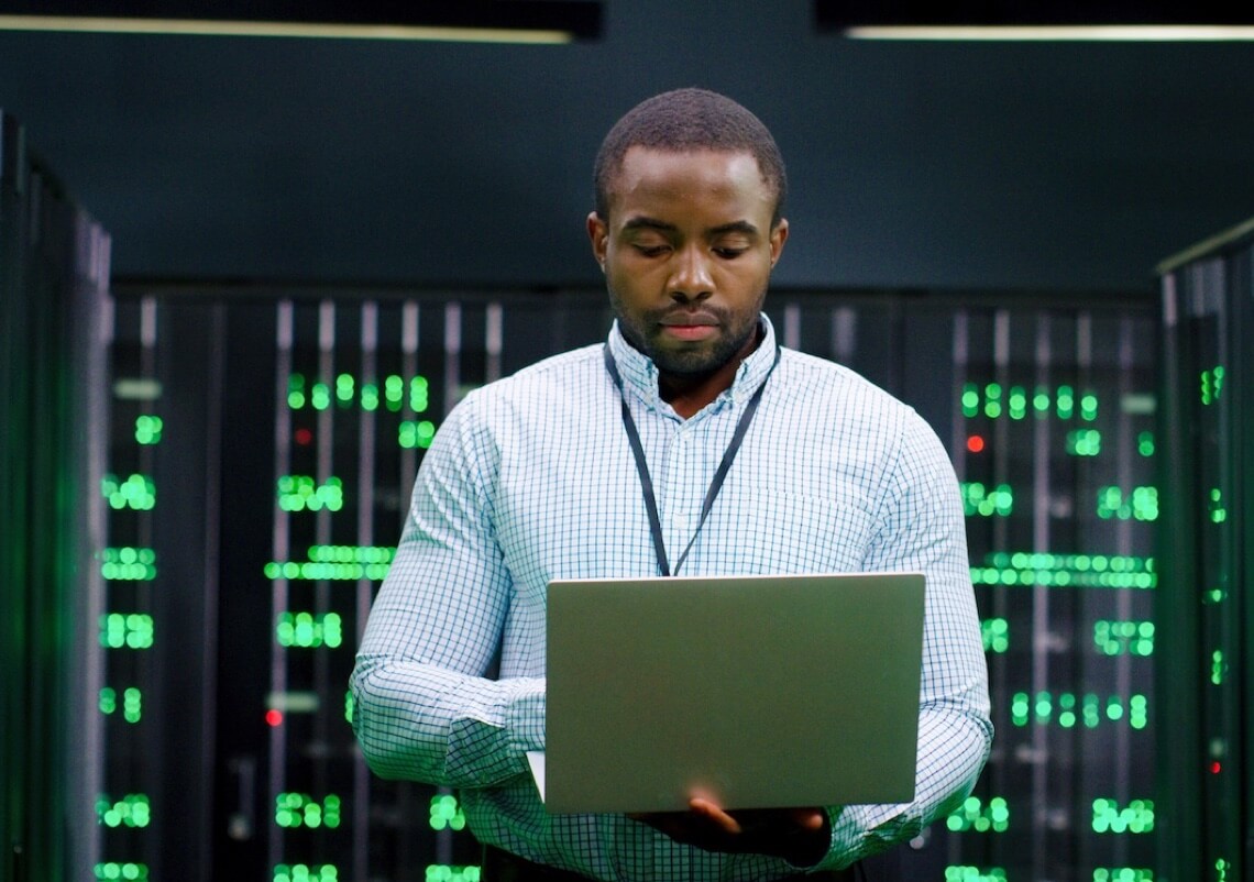 A systems administrator standing in front of servers while working on a laptop.