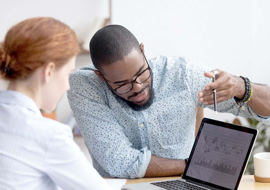 A young black male IT analyst explains something on a computer to a redheaded female colleague.