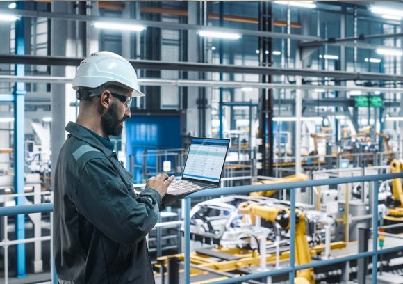 A young male supply chain manager is checking inventory on a laptop on a factor floor. He has a beard and is wearing a hard hat.