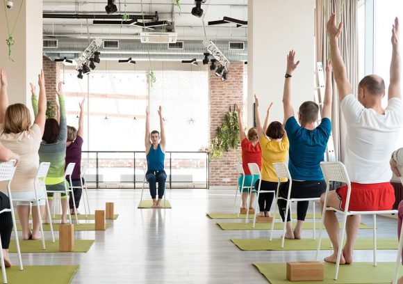 Young male group fitness instructor leading a seated yoga class in a large bright room.