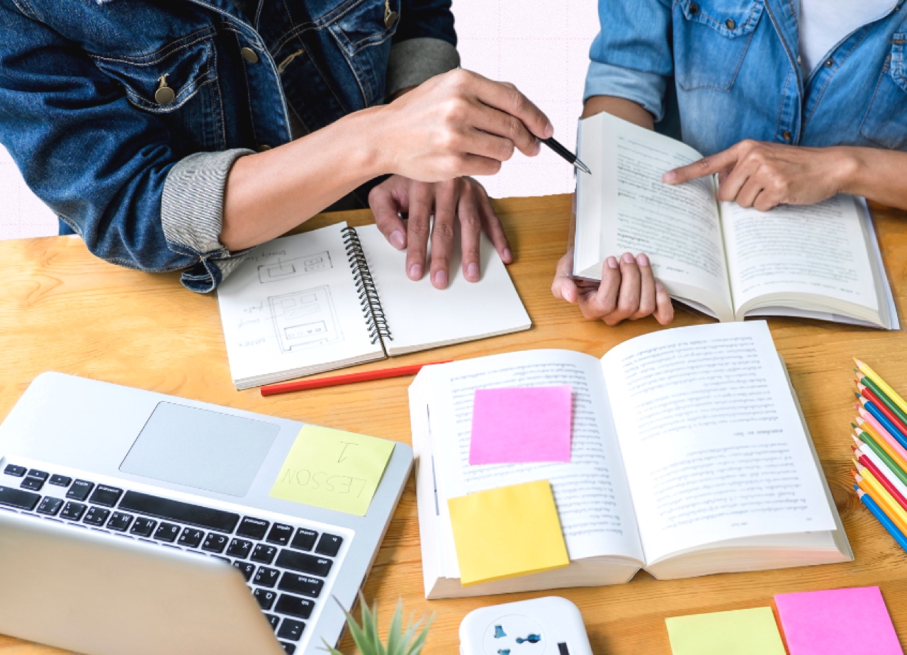 Close up of desk with two people studying using multiple books, pencils and post it notes.