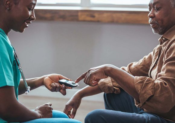 A young Black female nurse checking the glucose level of an older Black patient while both are seated.