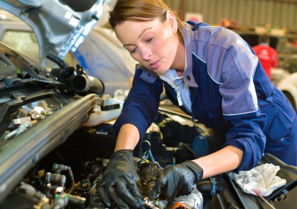 A young, white female auto mechanic works under the hood of a car on the engine.
