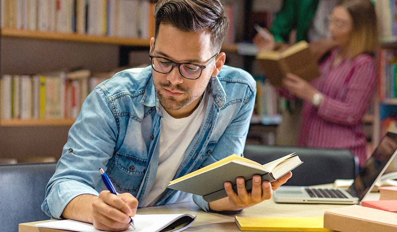 A young white male wearing glasses and a denim shirt studies and takes notes at a table while holding a book.