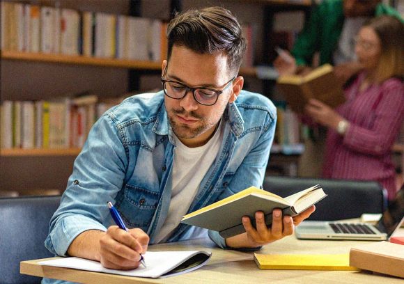 A young white male wearing glasses and a denim shirt studies and takes notes at a table while holding a book.