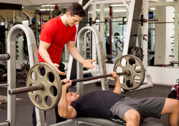 A young white male personal trainer spots another man on the bench press.