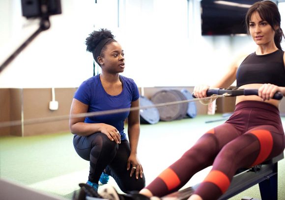 A young Black female personal trainer works with a female client on a row machine.