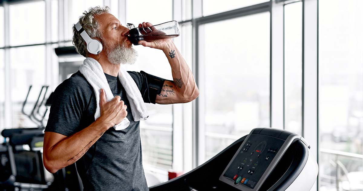 Older white male with beard and headphones drinking water while on a treadmill.