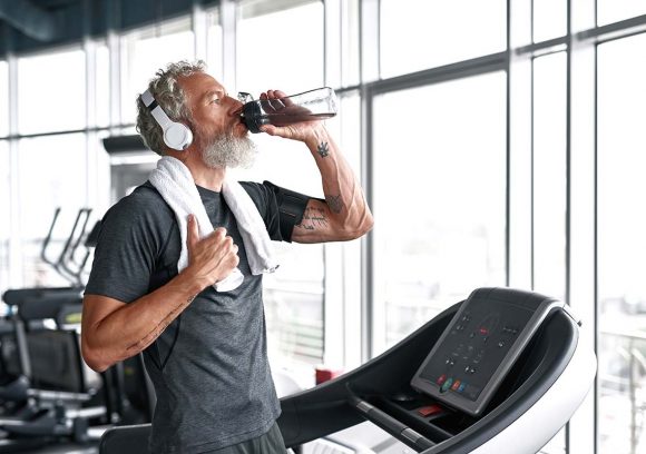 Older white male with beard and headphones drinking water while on a treadmill.