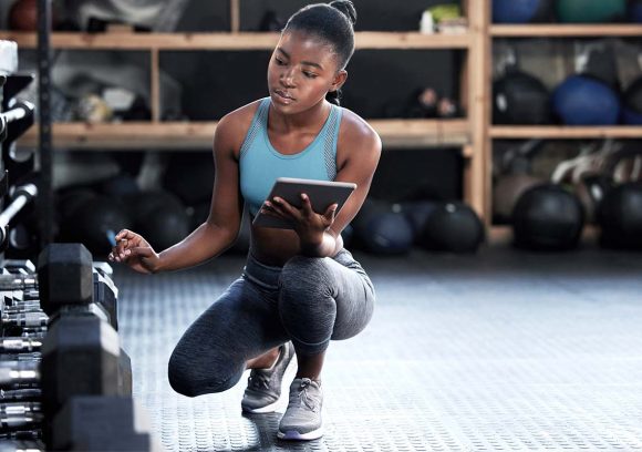 A young Black woman in athletic wear looks at a tablet while crouching next to a weight rack.