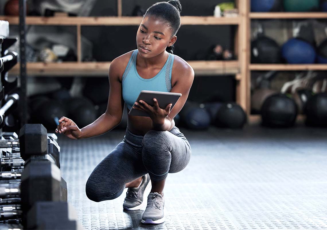 A young Black woman in athletic wear looks at a tablet while crouching next to a weight rack.