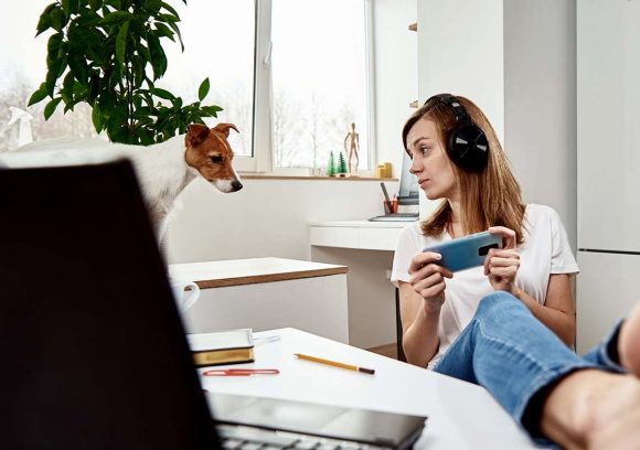 Young white woman wearing headphones is distracted while trying to study holding a phone and looking at a small dog with her feet up.
