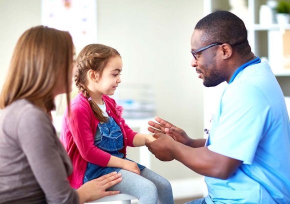 A Black family nurse practitioner checks a young female patient's hand while her mother looks on.