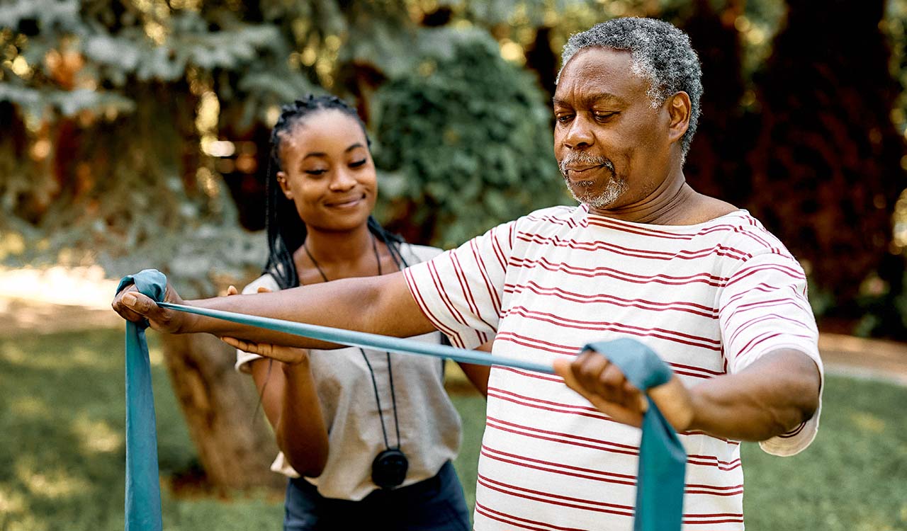 An older Black man using a resistance band outside under the guidance of a young Black female trainer.