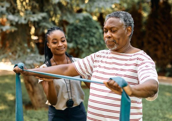 An older Black man using a resistance band outside under the guidance of a young Black female trainer.