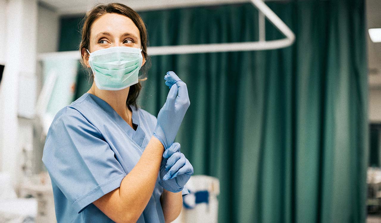 A female progressive care nurse wearing a mask looks to the side of a hospital room while putting on gloves.