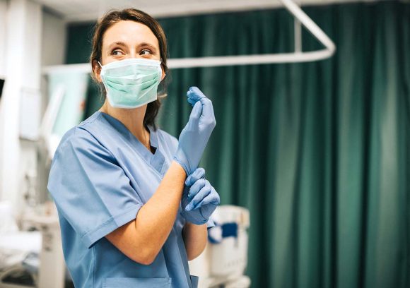 A female progressive care nurse wearing a mask looks to the side of a hospital room while putting on gloves.