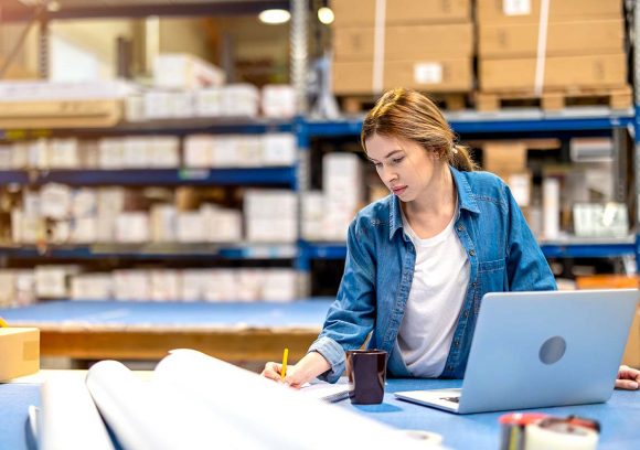 A young woman wearing a blue shirt working through supply chain logistics in a warehouse with a computer and coffee.