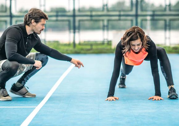 A young male fitness trainer coaches a young female client through dynamic stretches on an outdoor court.