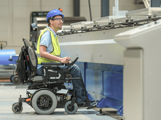 A worker with a muscular dystrophy in a wheelchair wearing a hard hat and safety vest operates machinery in an industrial setting.