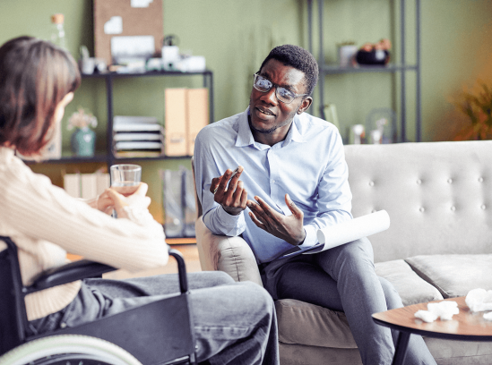 A certified rehabilitation counselor, holding a notebook, attentively listens to a client in a wheelchair in a cozy office.