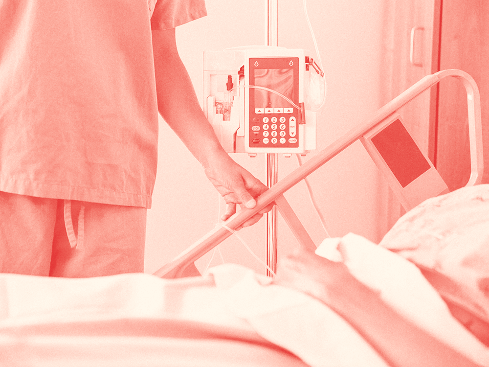 A nurse standing next to a patient's bed with an IV pump in the background.