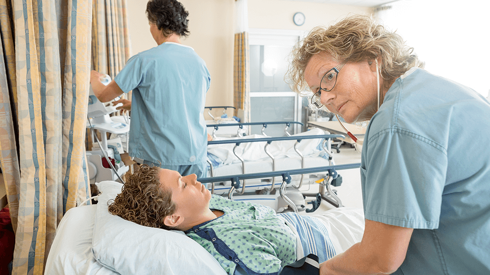A post-anesthesia nurse listens to a patient’s heartbeat in a hospital recovery room as the patient lies in bed recovering from an operation.