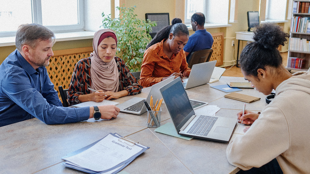 A group of students sitting with their instructor at a table taking notes from their computer.