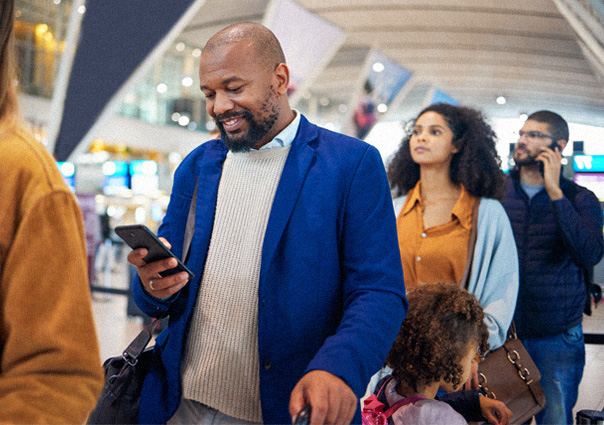 A man standing in a line of people at the airport looking down at his phone while smiling.